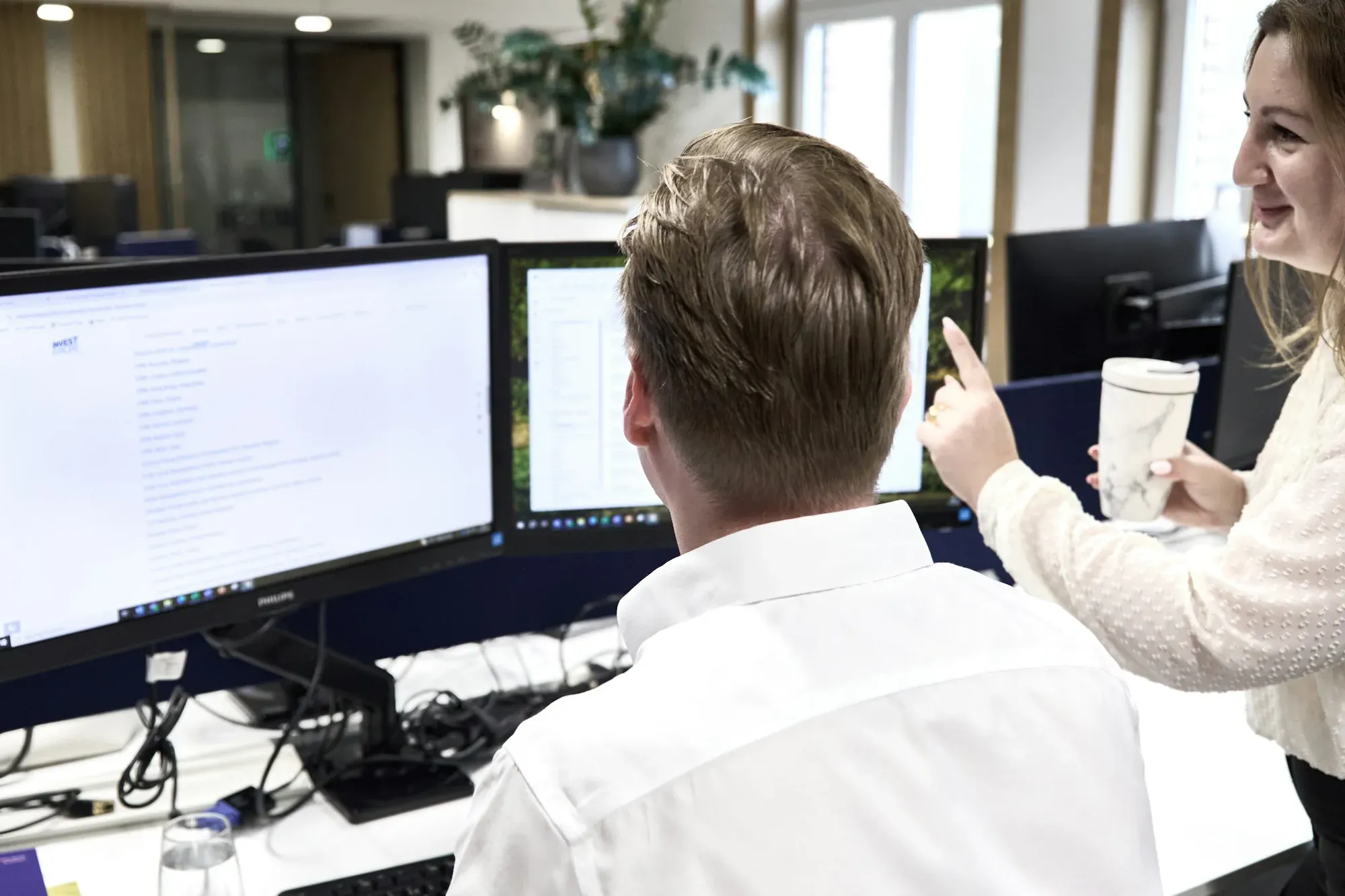 Zwei Kollegen arbeiten im Büro an zwei Bildschirmen, eine Kollegin zeigt auf einen Eintrag